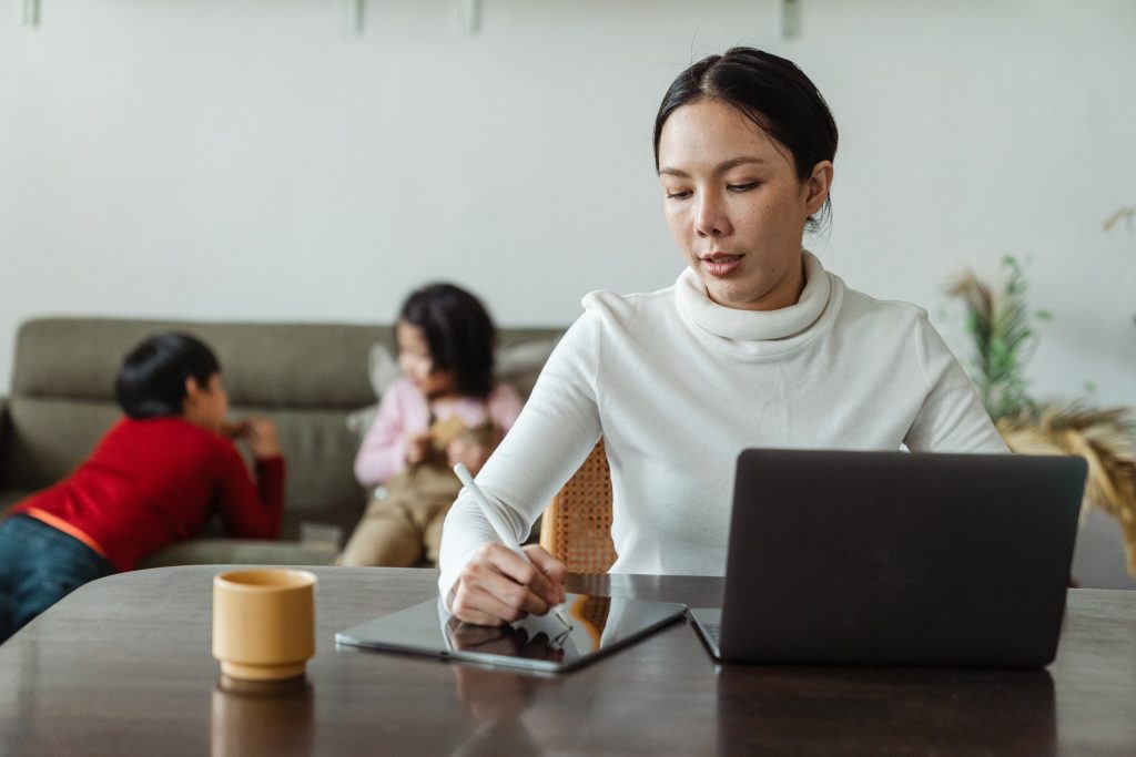 Madre trabajadora de origen étnico que utiliza una tableta y un portátil en casa, con los niños jugando de fondo.