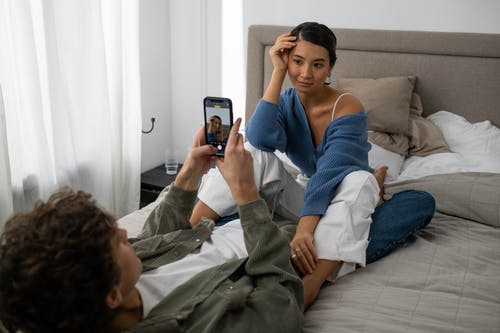 Young man taking a photo of a young woman sitting on the bed. 