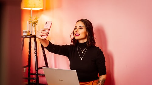 Mujer sentada delante de un portátil, haciéndose un selfie y escribiendo un pie de foto para el selfie en Instagram