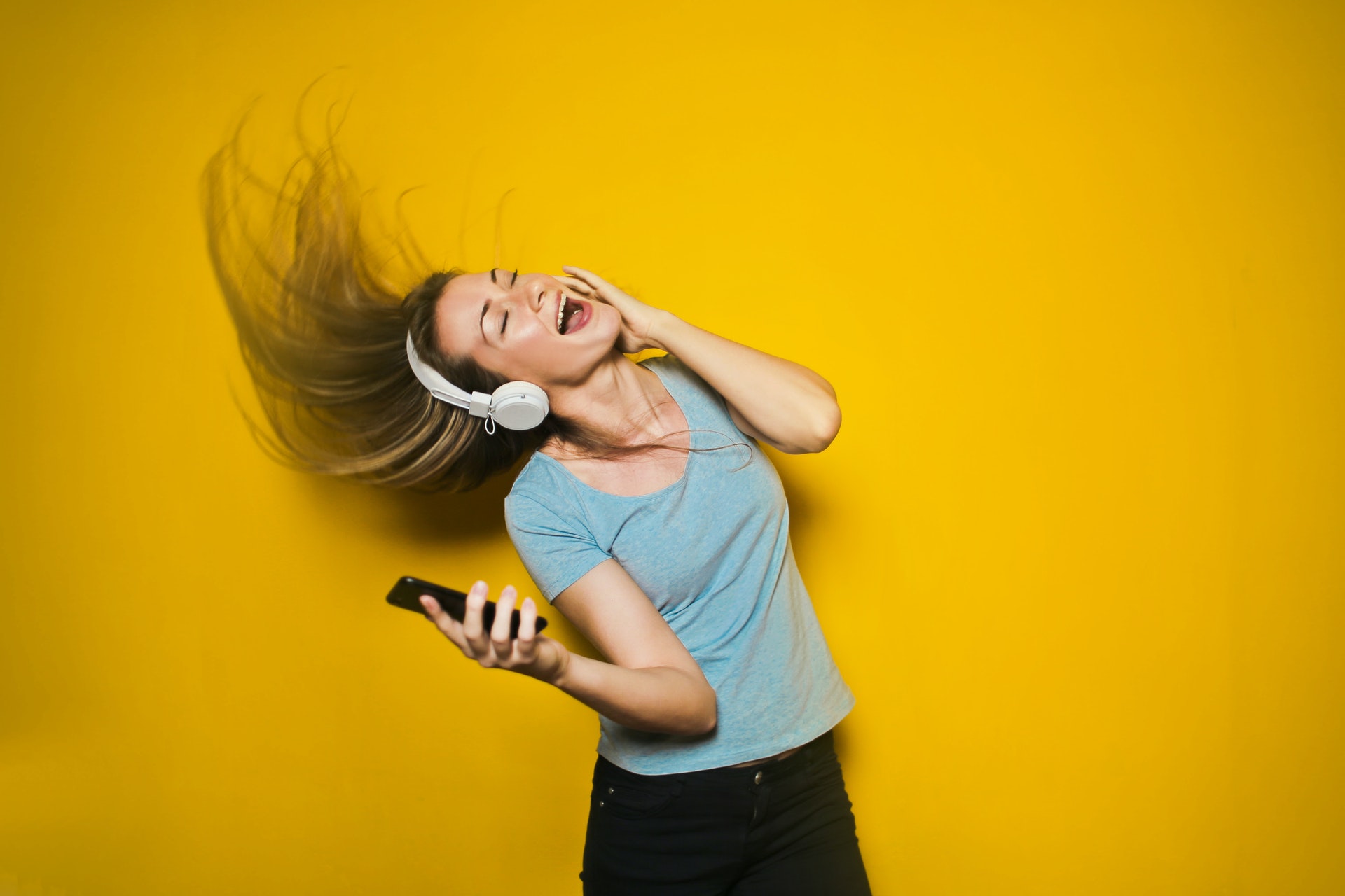黄色い背景の壁紙に隔離された、髪を動かしながらヘッドホンとスマートフォンで音楽を聴き、踊る興奮した女性。