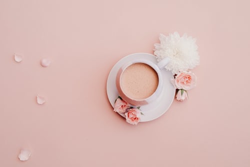 An Instagram picture using a pastel pink filter featuring a white cup filled with liquid and sitting on a white saucer. 