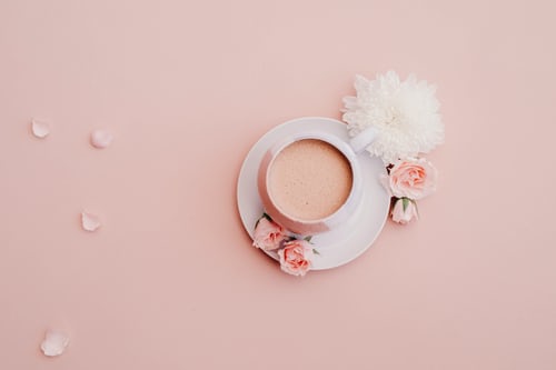 Una foto de Instagram con un filtro rosa pastel en la que aparece una taza blanca llena de líquido y sentada sobre un platillo blanco. 