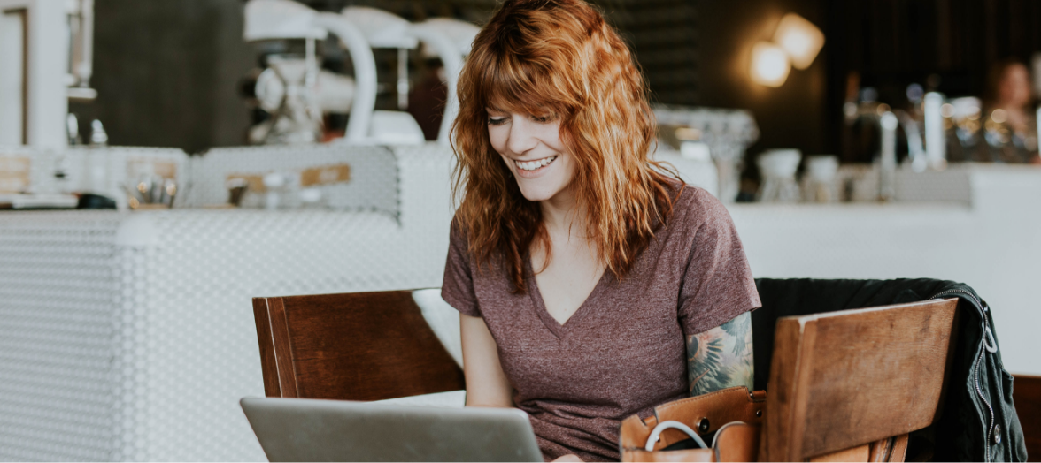A smiling woman with red hair using a laptop at a cafe table.