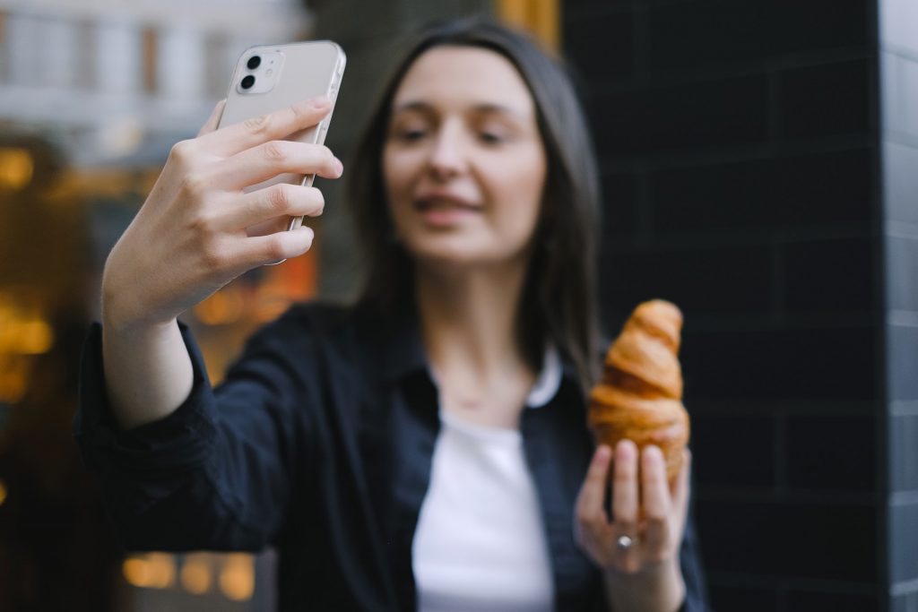 Woman taking a selfie with a smartphone while holding a pastry.