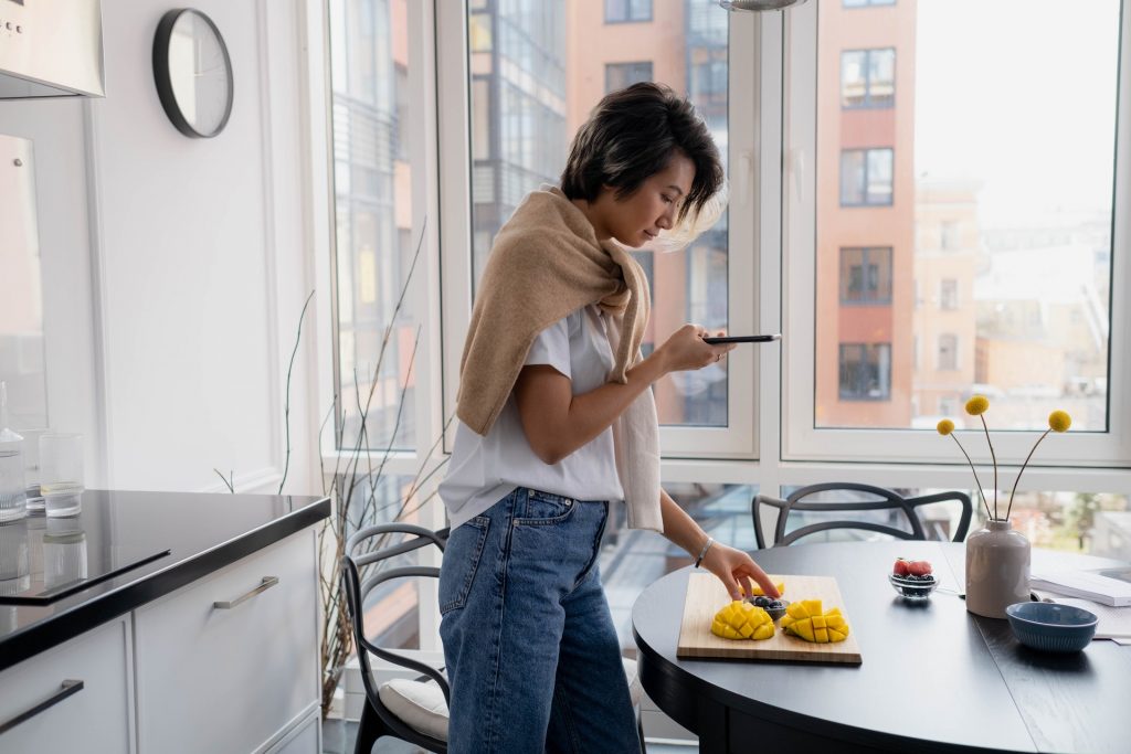 Woman standing in kitchen and using smartphone to learn how to make a reel on Instagram.