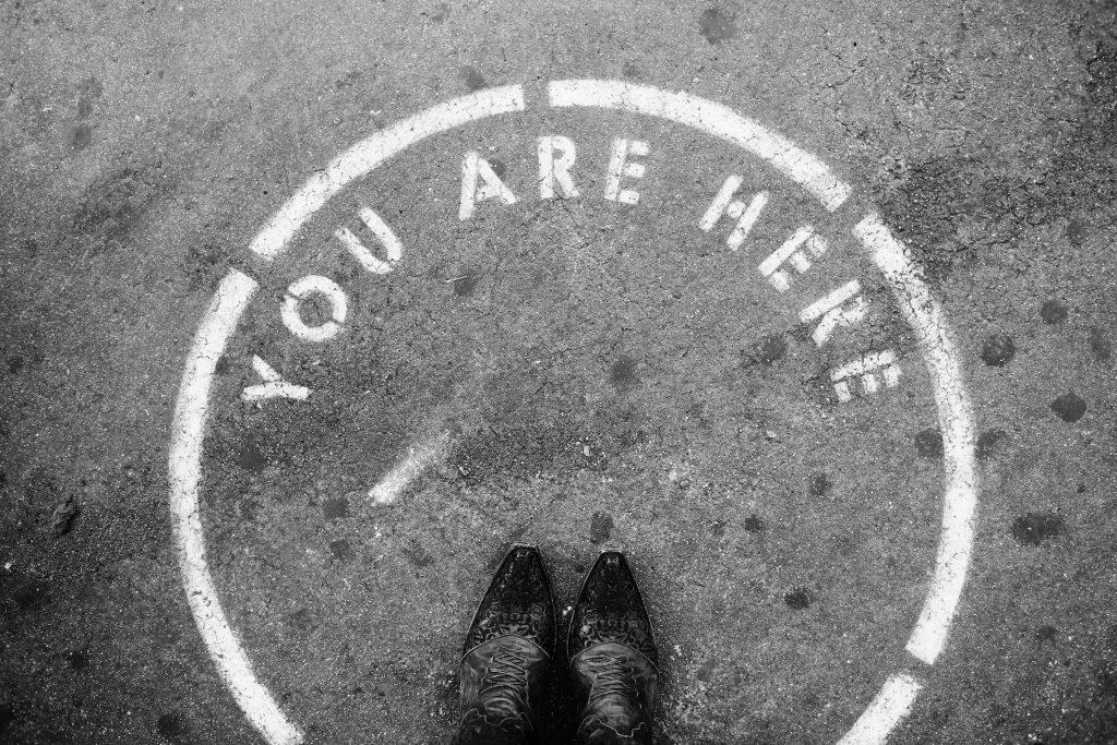 Wit geschilderde cirkel op beton met de woorden "U bent hier" en een paar schoenen op de voorgrond om locatiemarkering te demonstreren op Instagram. 