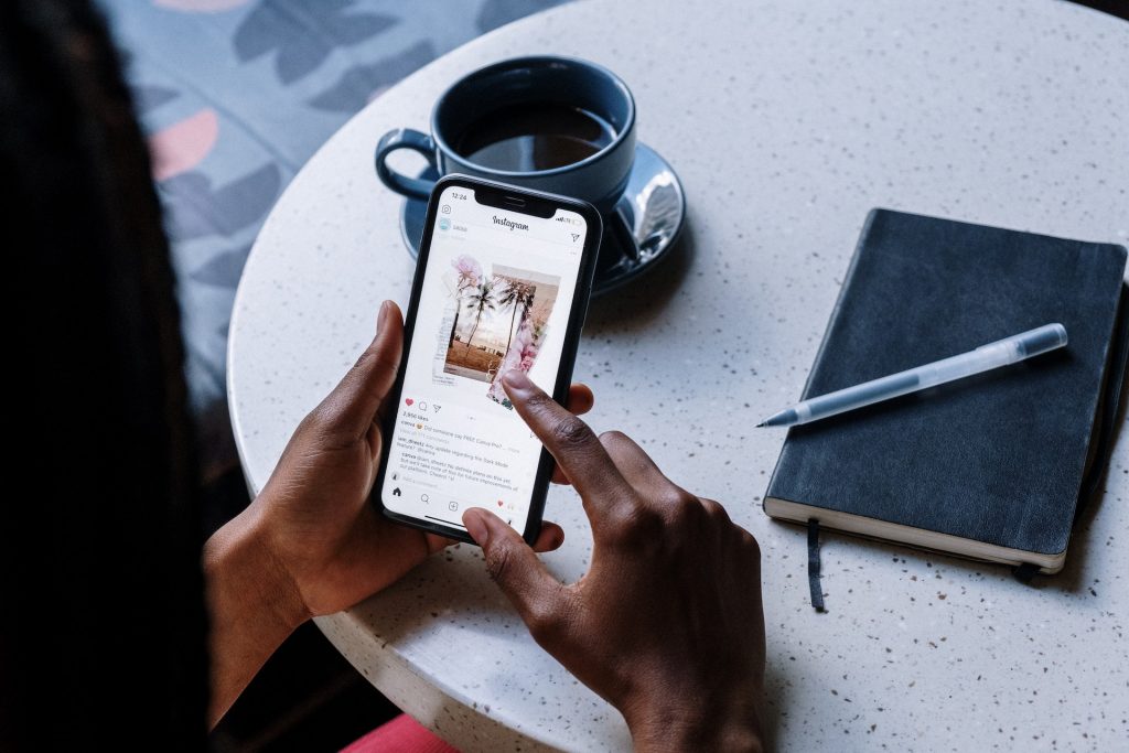 Hands holding black phone with instagram post on white table with a cup of coffee and a notebook with pen.