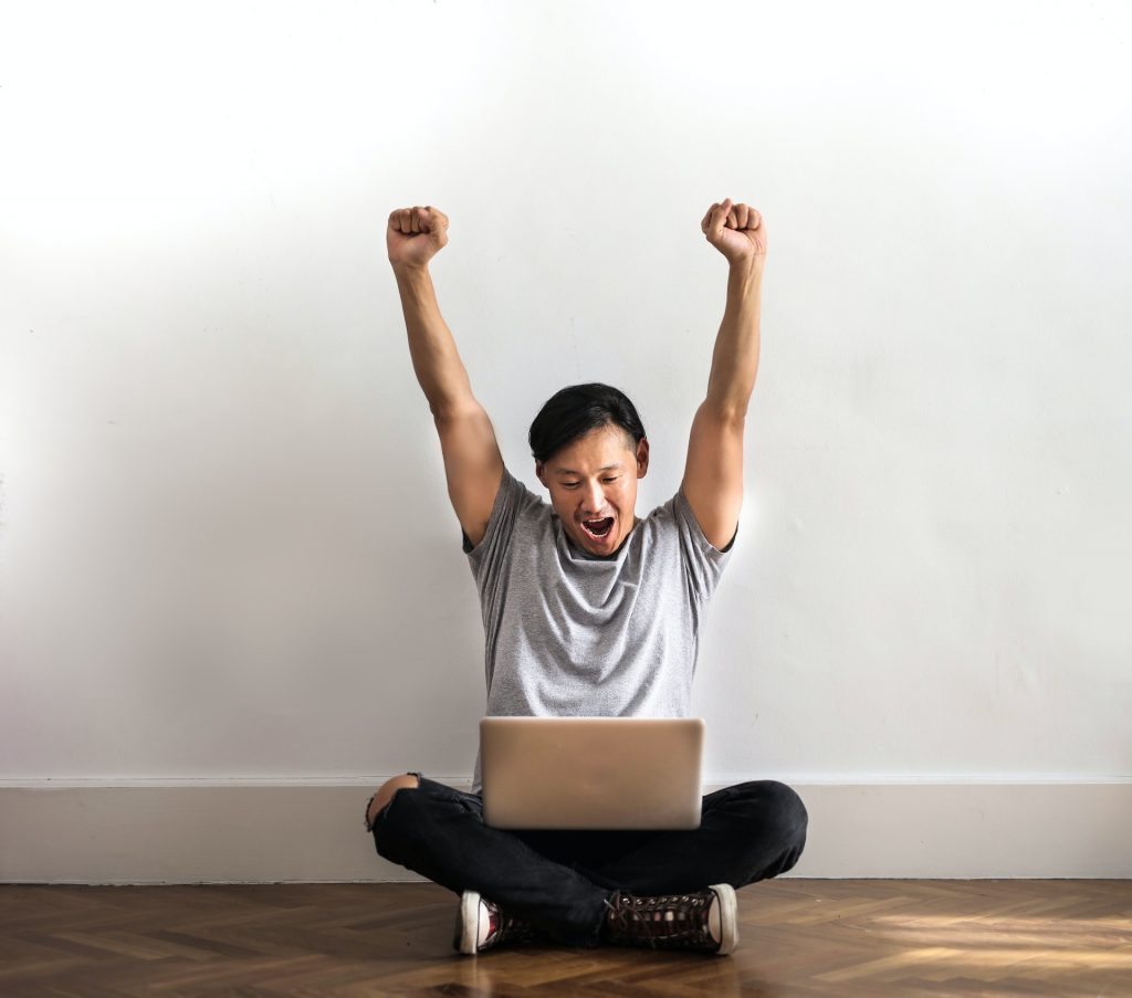 A man sitting on wooden floor and working on his laptop celebrating with his hands raised.