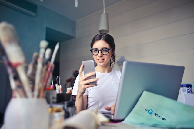 Uma mulher a sorrir enquanto segura um telemóvel em frente ao seu computador portátil.