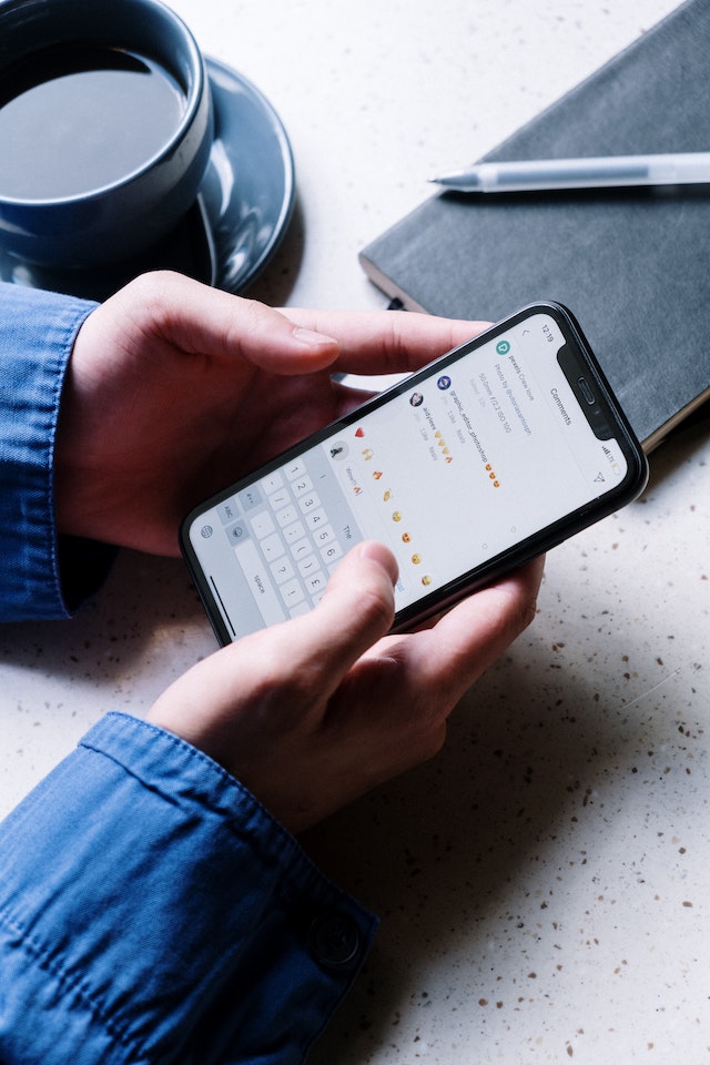 Person holding smart phone open to Instagram app on a table with coffee cup, notebook and pen.