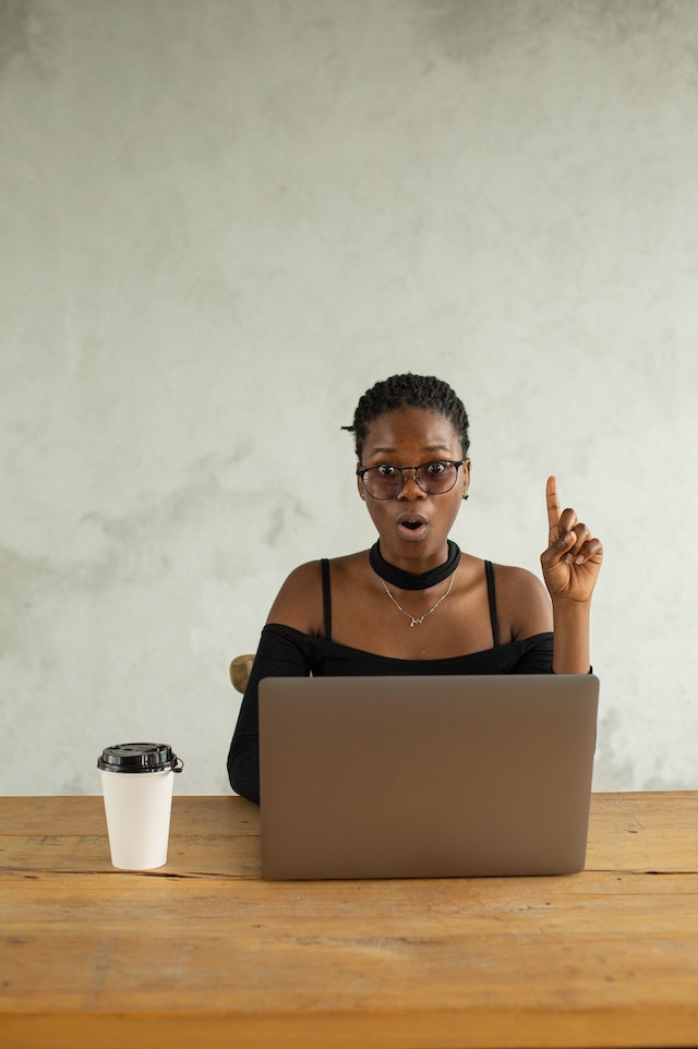 Een vrouw zit voor de laptop, haar vinger wijst omhoog, met een kop koffie naast haar. 