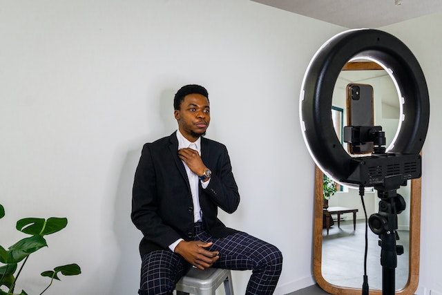Man modeling for a smartphone camera and ring light, with mirror and plant in the background.