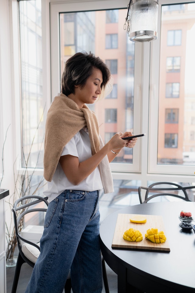 Frau steht in der Küche und benutzt ihr Smartphone, um zu lernen, wie man eine Rolle auf Instagram macht.