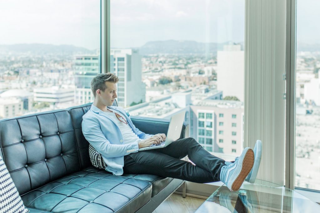 austin-distel-VvAcrVa56fc-unsplash | Path Social A man sitting on a couch working on his laptop with a view of a city in the background.