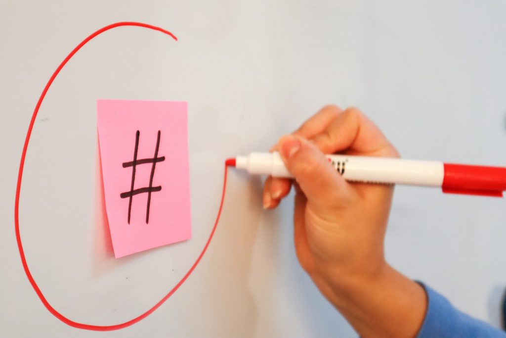 walls-io-iOnN7Et1FhA-unsplash | Path Social A hand drawing a red circle around a hashtag symbol on a pink Post-it note on a whiteboard.
