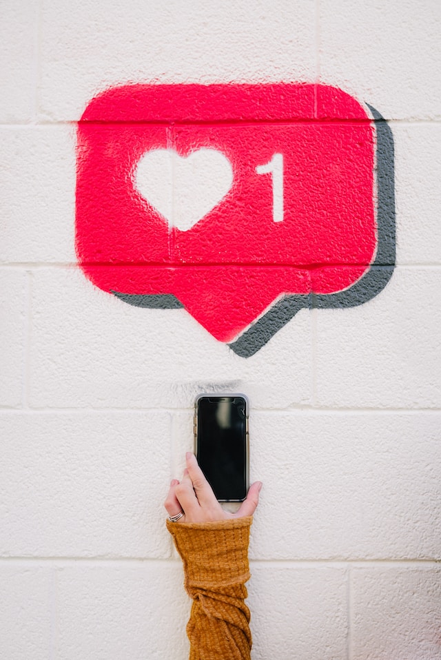 Una mano sosteniendo un teléfono con un globo de diálogo rojo encima con un corazón y el número 1 indicando hashtags en Instagram ganando likes.