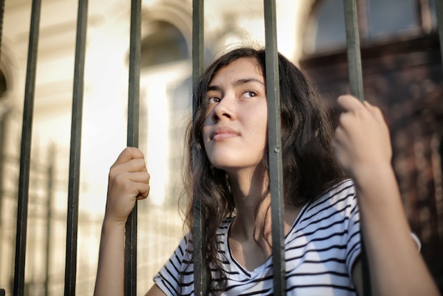 Woman looks through metal prison bars with her hands tightly gripping these bars