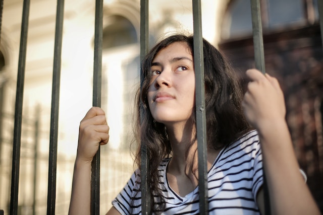 Woman looks through metal prison bars with her hands tightly gripping these bars.