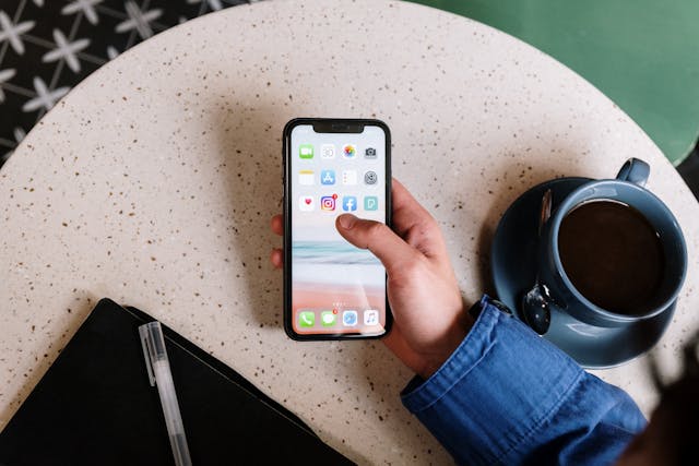 pexels-cottonbro-5053738 | Path Social Uma pessoa segura um iPhone preto por cima de uma mesa de café com aplicações de redes sociais no ecrã.