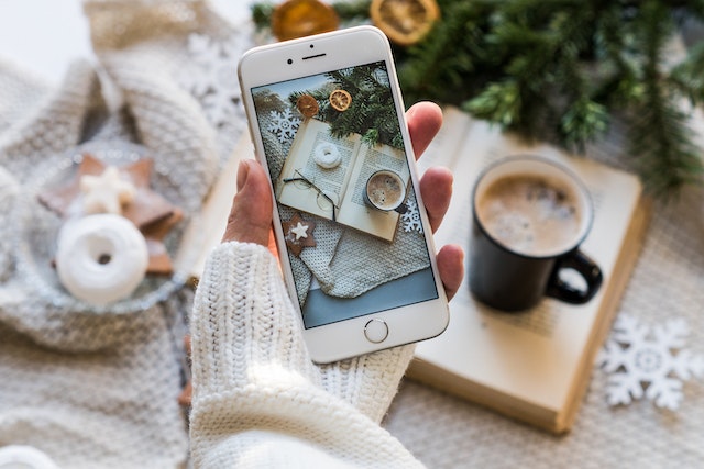  Personne en pull douillet prenant une photo d'un livre, de lunettes et d'une tasse de café chaud.