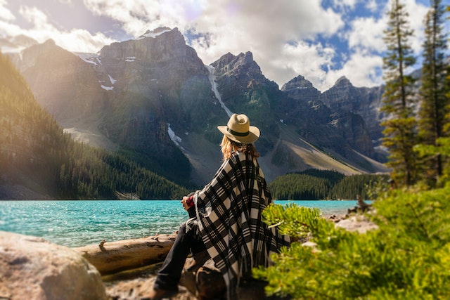 Uma mulher com um chapéu senta-se numa rocha com vista para um lago e para as montanhas, enrolada num cobertor.