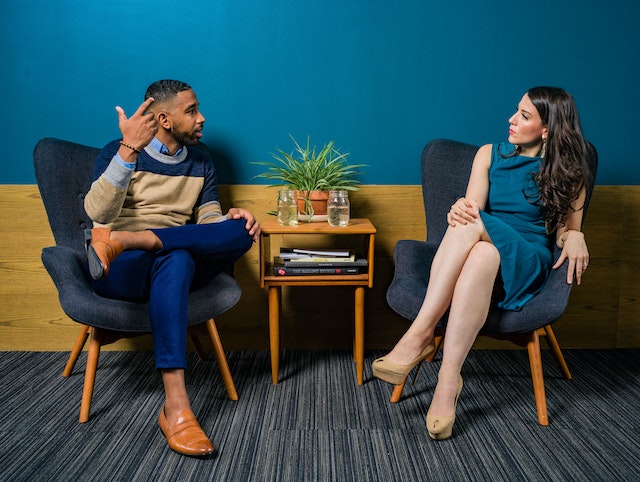 Woman Wearing Teal Dress Sitting on Chair Talking to Man
