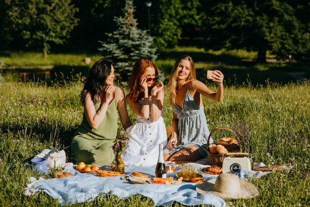A woman holding her phone while taking a photo with her two friends sitting outside on a picnic blanket.