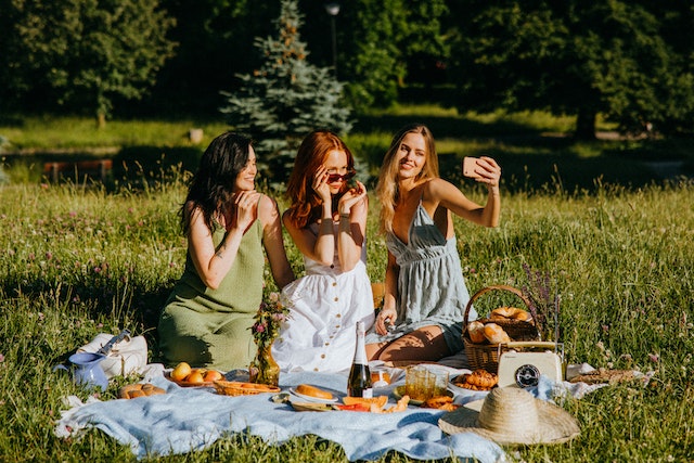 Una mujer sostiene su teléfono mientras hace una foto con sus dos amigas sentadas al aire libre en una manta de picnic.