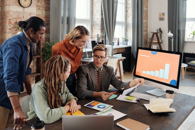 A Group of People Discussing Beside a Desktop with Graph on Screen 