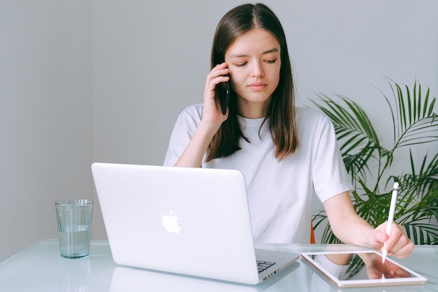 Woman in a white crew neck T-shirt using a silver Macbook while on the phone and writing in a notebook.