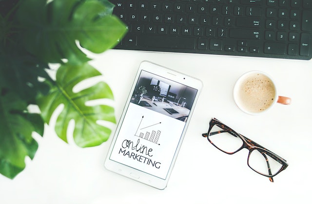 Brown-framed eyeglasses next to coffee, an iPad, a keyboard and a house plant.