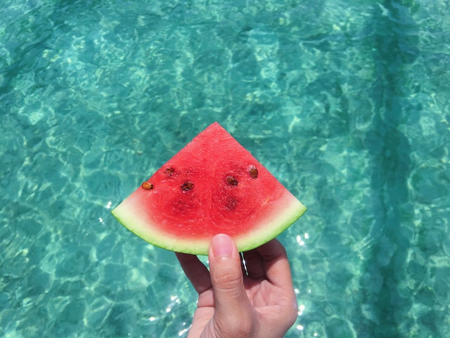 Unseen person’s hand holding a slice of watermelon over a swimming pool.