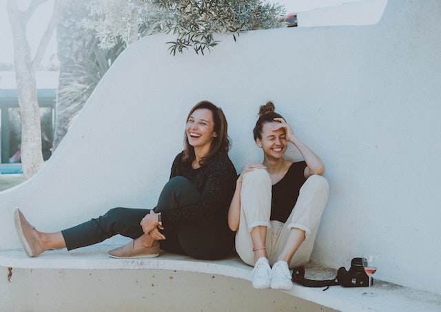 Twee vrouwen zitten op een wit bankje te lachen.