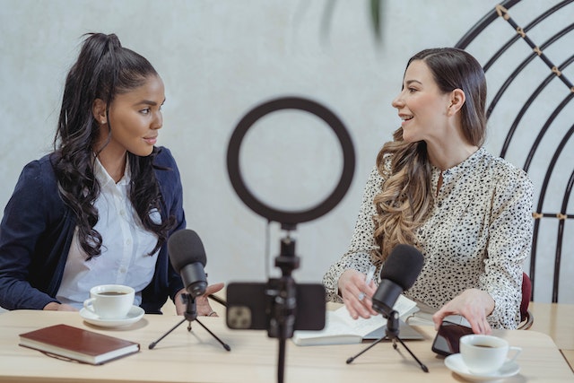 Deux femmes assises devant des micros enregistrent une interview avec des carnets et des tasses de café.