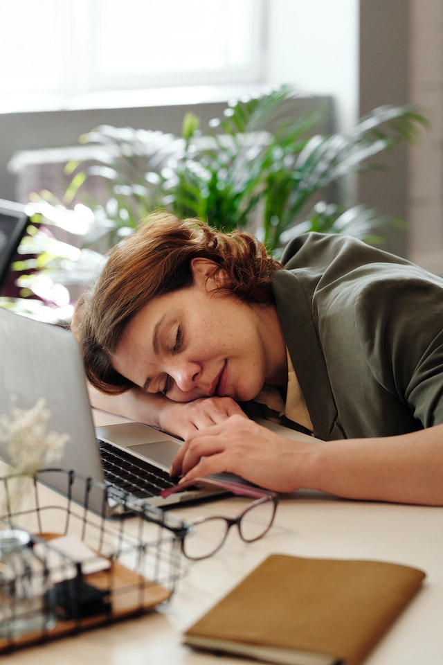 Mulher deitada de cabeça para baixo na secretária a dormir ao lado do seu computador portátil.