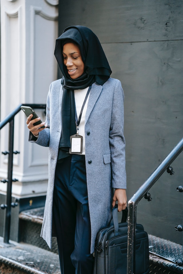 Happy Muslim black businesswoman surfing smartphone on building stairs.