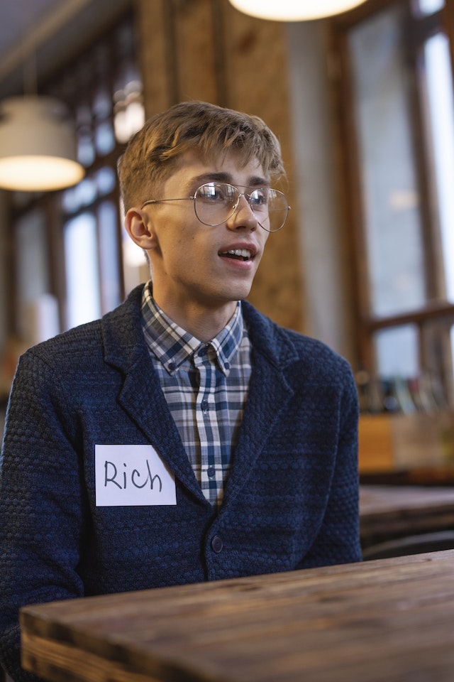 Uomo con cartellino e camicia a quadri blu e maglione con occhiali da vista.