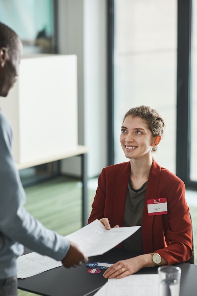 Femme assise à une table, vêtue d'un blazer orange et portant un badge, souriant à une autre personne.