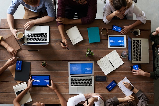 Collega's zitten rond een houten tafel tijdens een vergadering met computers en mobiele apparaten.