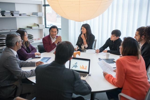 Oito pessoas sentadas numa mesa redonda durante uma reunião de trabalho.