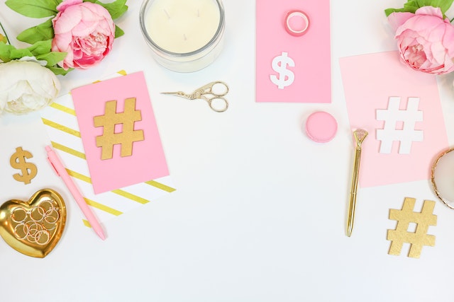 Image of paper hashtags and dollar signs surrounded by pink and white flowers and office supplies.