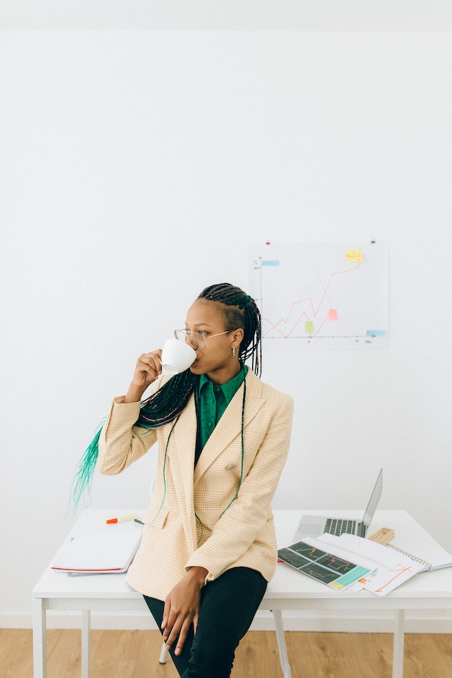 Woman in a beige coat drinking from a white cup in front of her work desk.