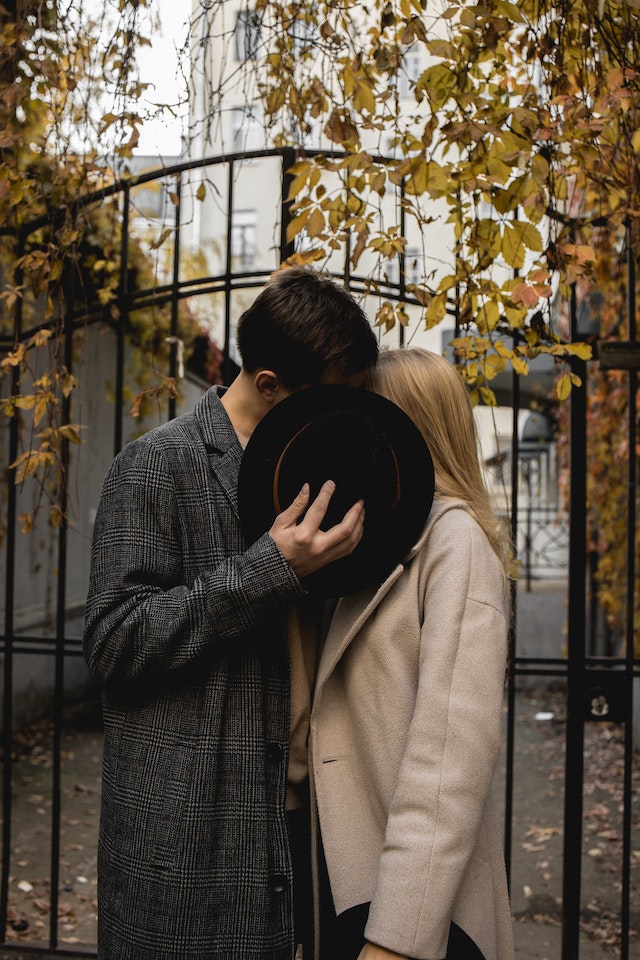 Casal a beijar-se atrás de um chapéu, diante de um portão de metal e folhas de outono. 