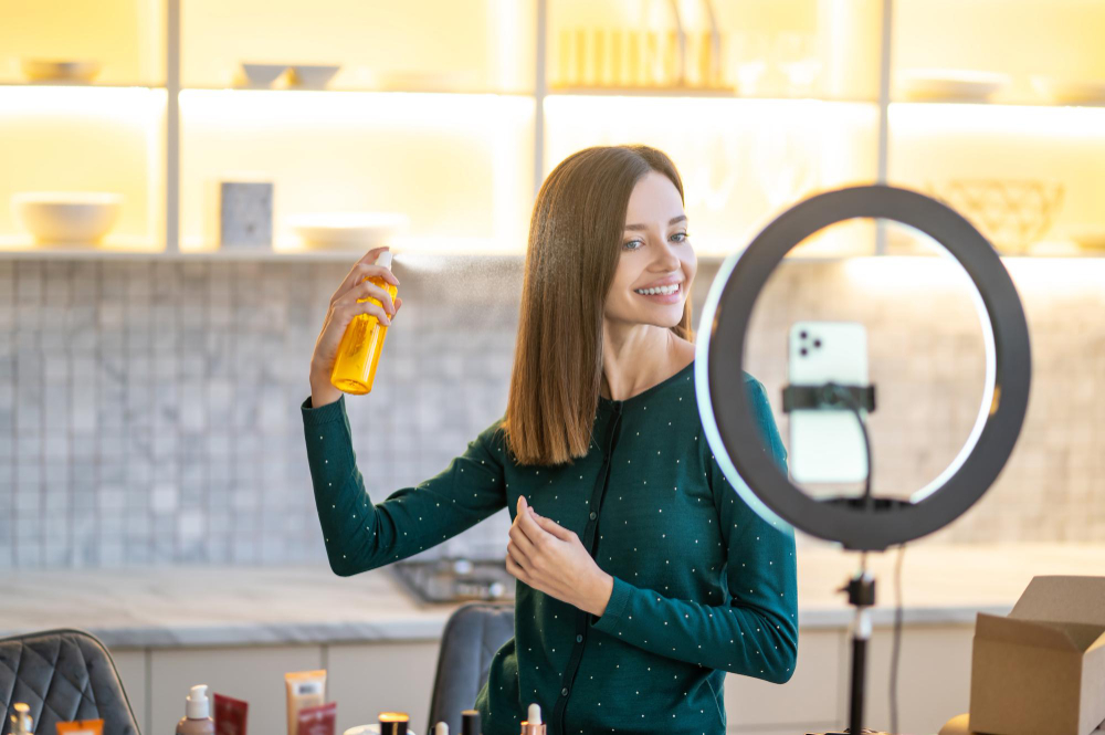 smiling-young-woman-presenting-hairspray-during-online-beauty-tutorial | Path Social une femme utilisant un flacon pulvérisateur pour se coiffer
