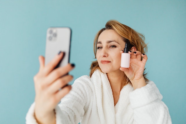 a woman in a white robe holding a small bottle of liquid and a phone