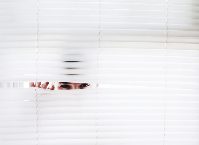 A person hiding and peeking behind blinds.