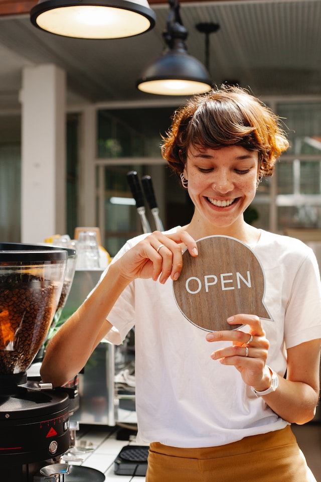 Une femme travaillant dans un café local tient une pancarte en bois indiquant "Ouvert".