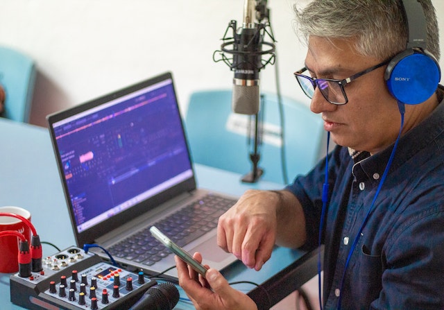 A podcaster records content using his smartphone, headphones, a mic and laptop.