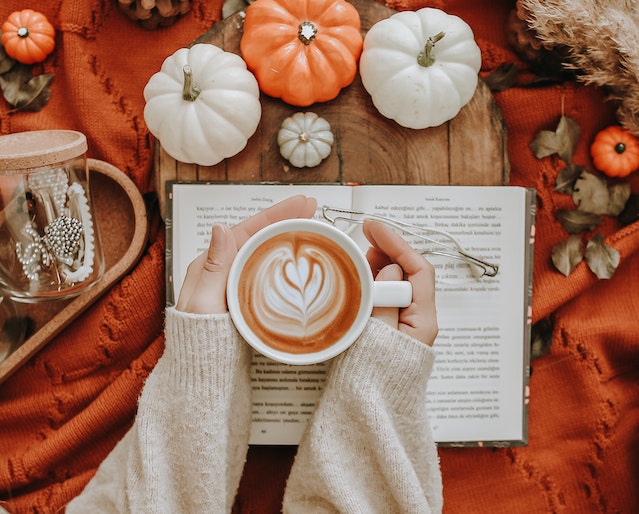 Frau mit Milchkaffee in der Hand, die auf einem mit Kürbissen bedeckten Tisch ein Buch liest.