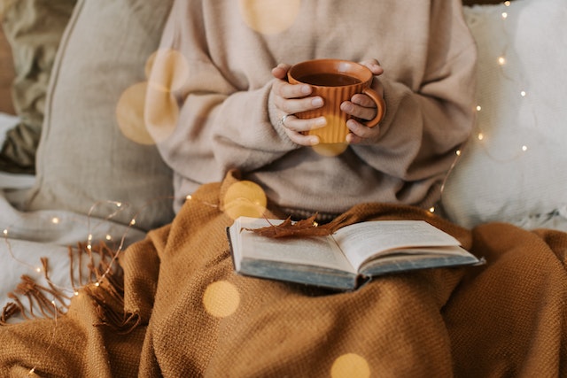 Femme vêtue d'un pull beige confortable lisant un livre en buvant un café.